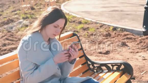 一幅美丽的黑发女人坐在公园的长凳上在手机里读和打信息的画像视频的预览图