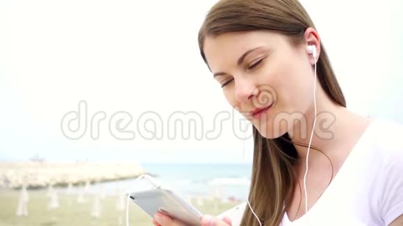 年轻微笑的女人独自在海滩上使用手机女性通过手机慢动作听音乐视频的预览图