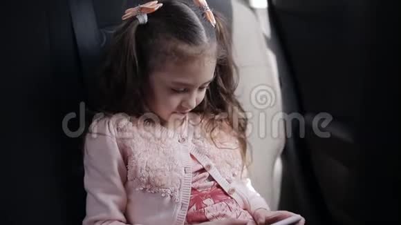 漂亮的蓝眼睛小女孩坐在后座的车里她把游戏集中在电话上视频的预览图