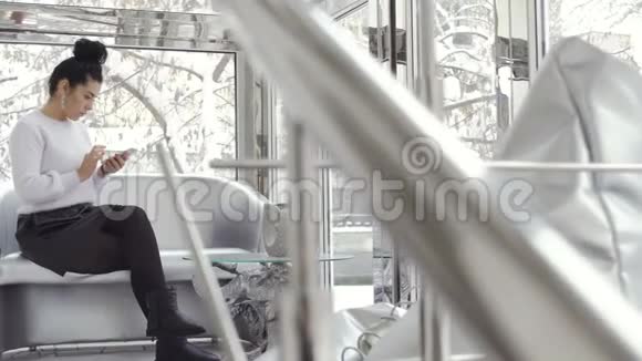美丽的黑发女人在靠近窗户的沙发上用电话放松视频的预览图