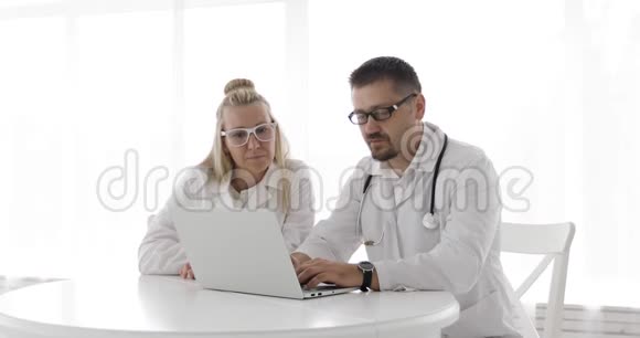 两个穿着医用长袍的医生坐在一张白色的桌子上用笔记本电脑视频的预览图