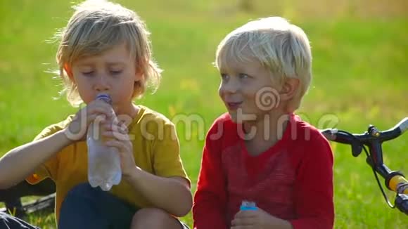 两个小男孩骑自行车坐在草坪上喝塑料瓶里的水的慢动作镜头淡水概念视频的预览图