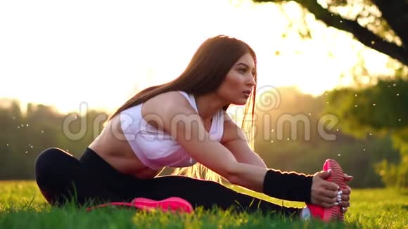 阳光下的公园里一个穿着粉红色鞋子的运动型深色头发的女孩在日落时慢动作伸展肌肉视频的预览图