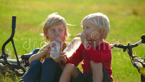 两个小男孩骑自行车的慢镜头坐在草坪上从塑料瓶里喝水淡水概念视频的预览图