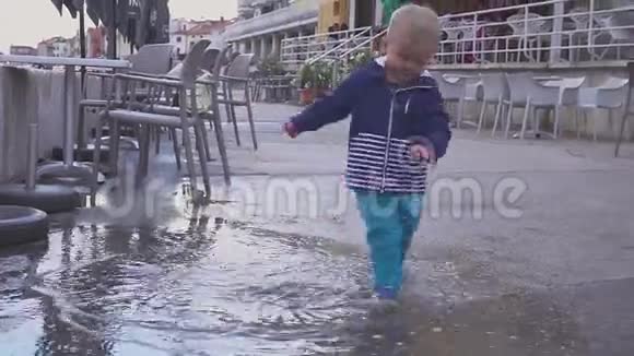 这个男孩在雨中的水坑里玩他在雨中快乐两岁的男孩在人行道上奔跑概念视频的预览图