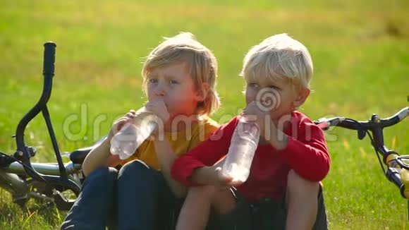 两个小男孩骑自行车坐在草坪上喝塑料瓶里的水的慢动作镜头淡水概念视频的预览图