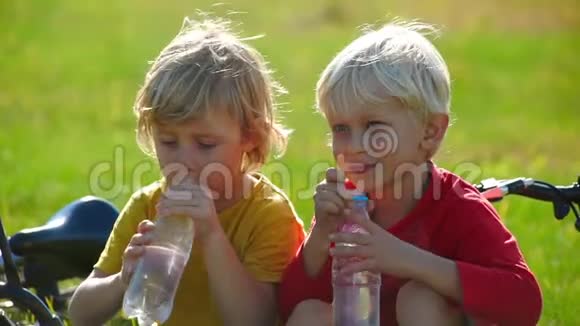 两个小男孩骑自行车的慢镜头坐在草坪上从塑料瓶里喝水淡水概念视频的预览图