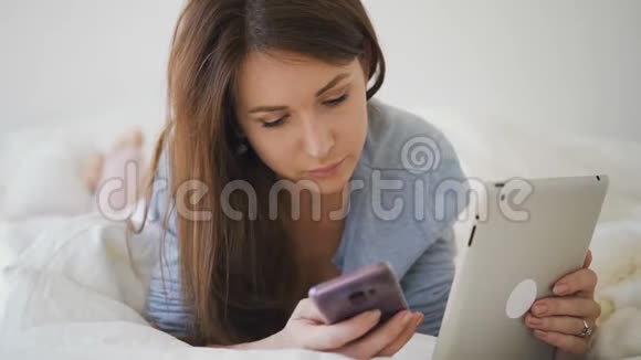 美丽的女人躺在床上在卧室的内部使用智能手机和平板电脑视频的预览图