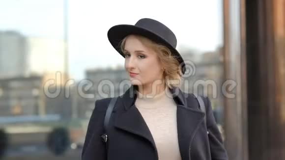 穿着黑色外套的贴身漂亮女孩戴着帽子带着耳机听音乐对抗模糊的现代建筑慢动作视频的预览图