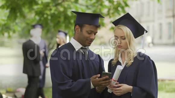 穿着毕业制服的年轻人看着手机屏幕讨论工作视频的预览图
