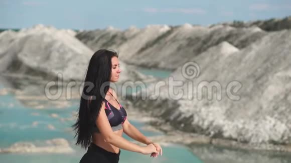 穿着黑色运动服的运动型黑发女孩在一个蓝水白垩沙场上训练在悬崖上做瑜伽视频的预览图