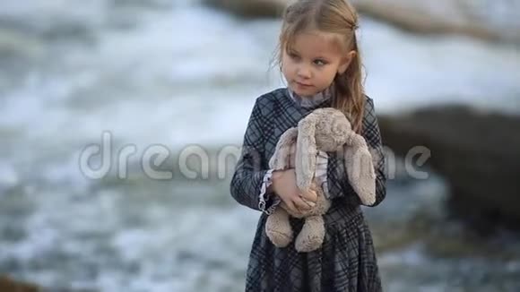 在一个刮风的日子里金发的小女孩站在海边女孩玩玩具兔子和软泰迪熊视频的预览图