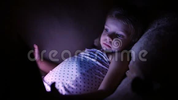 小可爱的女孩躺在沙发上在黑暗中玩平板电脑视频的预览图