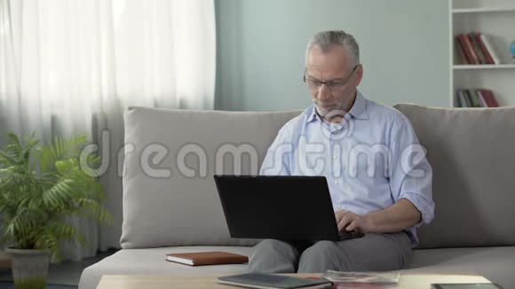 健康的老年男性坐在沙发上在交友网站笔记本电脑上观看照片视频的预览图