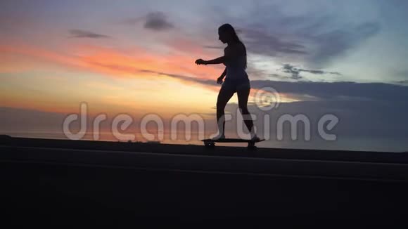美丽的女孩骑着滑板在夕阳的天空下上路视频的预览图