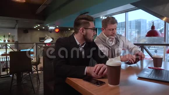 两个同事成年男子戴上眼镜和他年轻的同事在咖啡馆里讨论一个带笔记本电脑的项目视频的预览图
