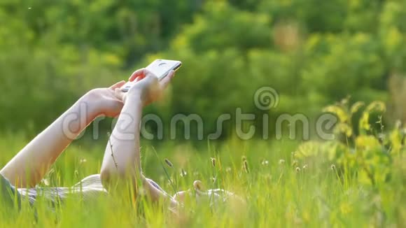 躺在绿色草坪上的快乐年轻女子在夕阳背景下的风景区使用智能手机视频的预览图