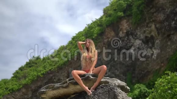 美丽的女人摆着相机坐在巴厘岛野生海滩上的岩石上视频的预览图