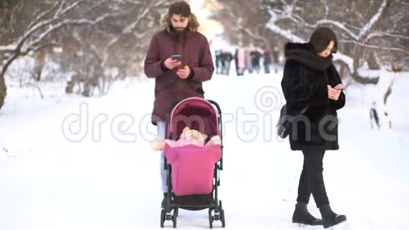 母亲和父亲沉迷于手机忽视婴儿车和冬季公园散步的婴儿家长忙着视频的预览图