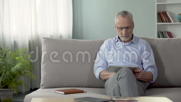 严肃的老人坐在沙发上在平板电脑上看新闻现代科技视频的预览图