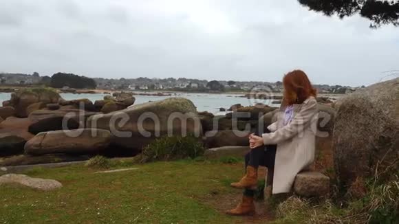 坐在长凳上的女孩在低潮时看着空荡荡的岩石海岸视频的预览图