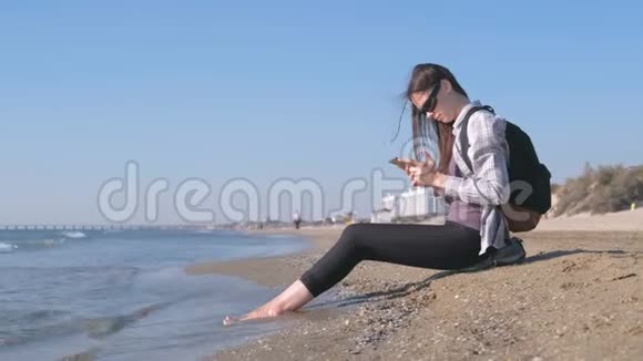 旅行者女孩带着背包坐在沙滩上在手机上输入一条信息视频的预览图
