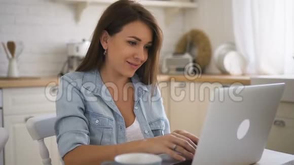 年轻漂亮的女人在家里的厨房里用笔记本电脑坐在桌子上视频的预览图