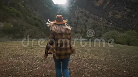 美丽的年轻女子旅行者在大自然中自由地穿越田野在群山把帽子扔到空中视频的预览图