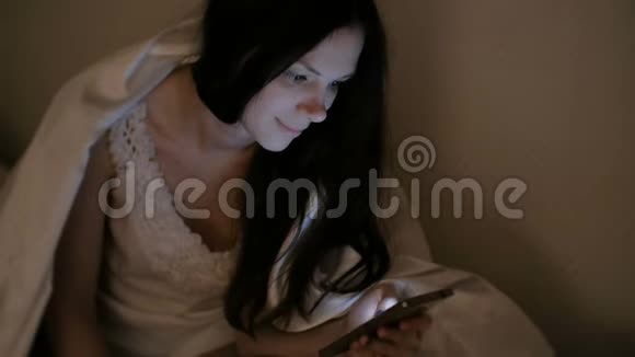 年轻漂亮的黑发女人在睡觉和微笑之前在床上看着手机里的社交网络视频的预览图