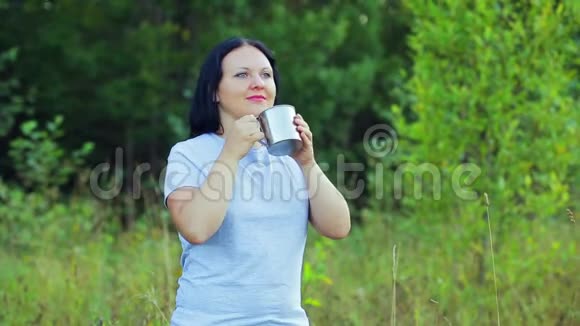 一个年轻的女人站在森林的边缘喝茶欣赏大自然视频的预览图