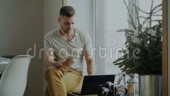 漂亮的年轻人坐在窗台上拿着笔记本电脑和相机在咖啡馆里浏览智能手机视频的预览图