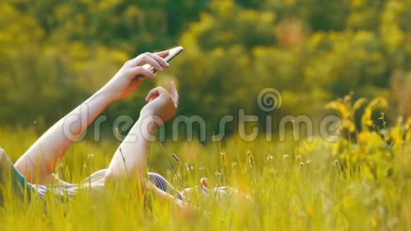 日落时分躺在草坪上的年轻幸福女子在风景名胜区使用智能手机视频的预览图