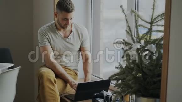 专注的男性摄影师带着相机和笔记本电脑坐在家的窗台上把她的照片分享给视频的预览图