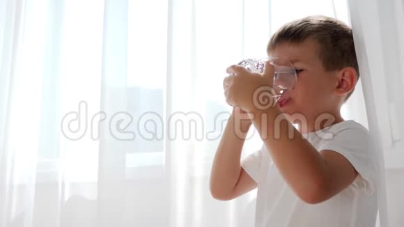 小男孩手里拿着一杯水在背景白色窗帘上喝矿泉水视频的预览图