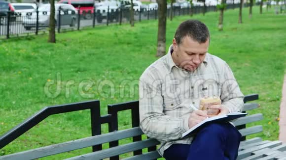 男人在公园长椅上吃快餐在笔记本上写东西视频的预览图