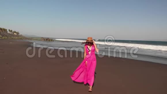 4巴厘岛黑色火山海滩上年轻女子穿着粉红色连衣裙行走的空中飞行镜头视频的预览图