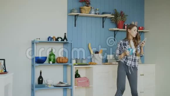 早晨快乐的女孩在厨房里跳舞唱歌在家里的智能手机上浏览社交媒体视频的预览图