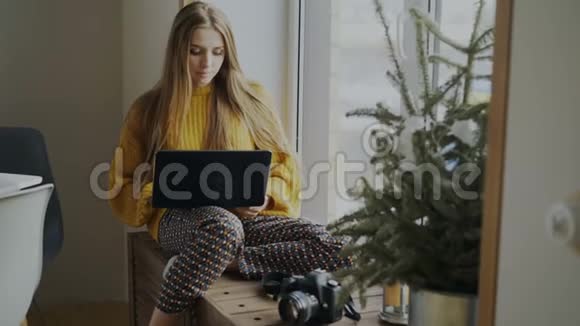 集中的女摄影师带着相机和笔记本电脑坐在家的窗台上把她的照片分享给视频的预览图