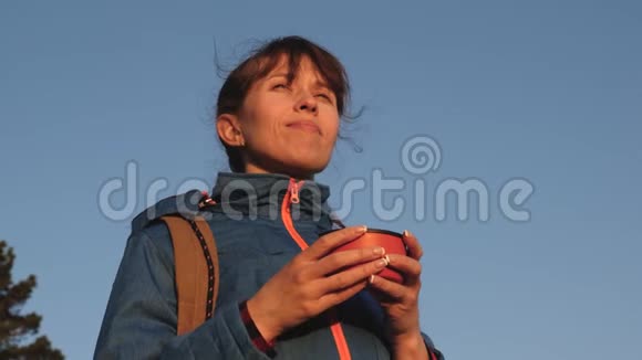 旅行者女人喝着热茶在蓝天上看日落女游客站在山顶喝酒视频的预览图