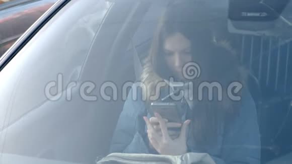 从汽车挡风玻璃上看到穿着蓝色羽绒服的年轻黑发女人看着电话视频的预览图
