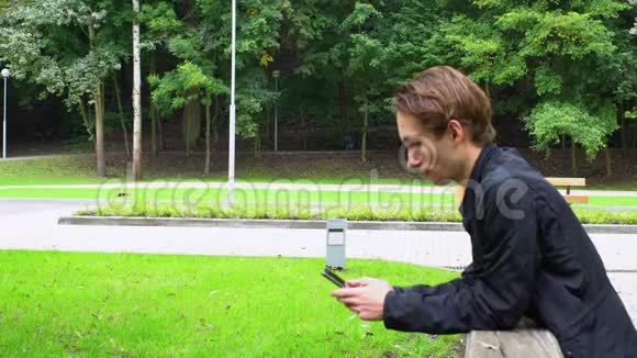 年轻人看着智能手机上的社交网络靠在公园的桥栏杆上视频的预览图