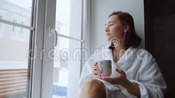 一个穿着白色外套的悲伤女人端着一杯茶坐在窗台上视频的预览图