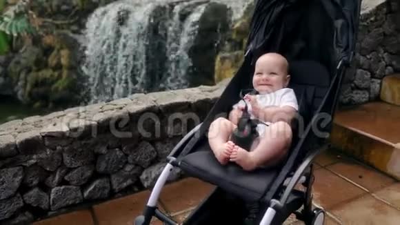 婴儿坐在瀑布附近的轮椅上笑着看着摄像机视频的预览图