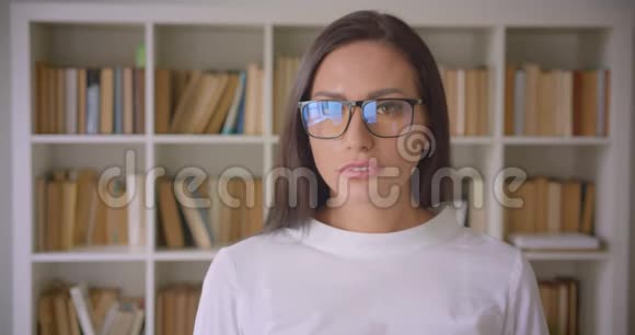 一位年轻漂亮的白种人女商人戴着眼镜背景上有书架的照相机的特写照片视频的预览图