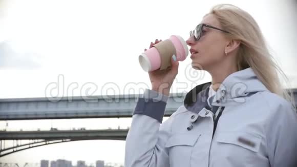 年轻漂亮的金发女郎喝着咖啡在河堤上散步慢动作视频的预览图