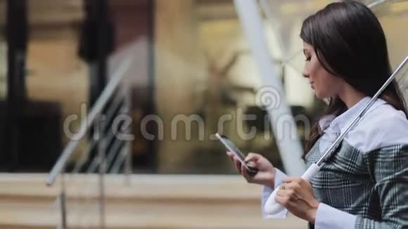 美丽的年轻商务女性在雨天用智能手机在街上行走微笑着拿着雨伞视频的预览图