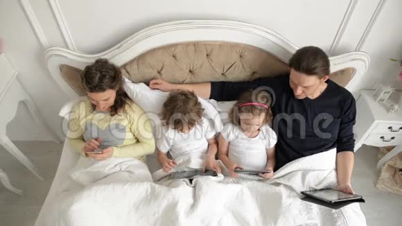 现代家庭使用移动设备两个孩子和他们的父母一起躺在床上度过周末视频的预览图