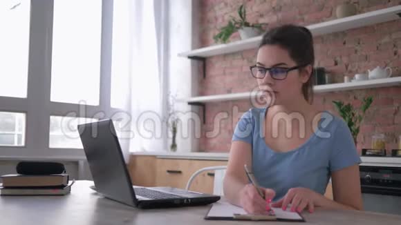 戴眼镜的学生用电脑笔记本电脑在线学习并在桌旁的剪贴板上写笔记视频的预览图