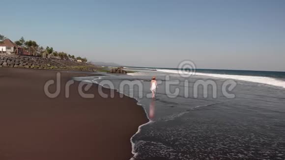 4巴厘岛黑色火山海滩上身穿白色礼服的年轻女子行走的空中飞行镜头视频的预览图