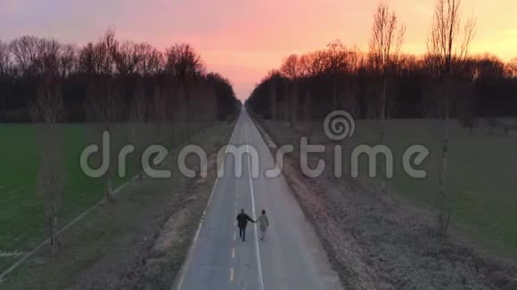 空中情侣牵着双手走在田野和林地之间的乡间小路上夕阳下五彩缤纷的浪漫天空视频的预览图